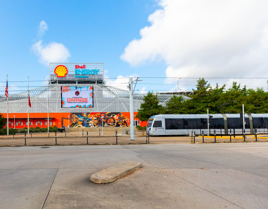 Front entrance of Shell Energy Stadium with a METRORail vehicle passing by.